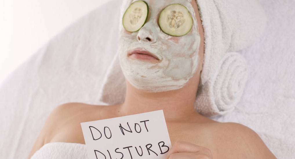 Young woman laying down on a massage bed with facial mask on and cucumber on the eyes and towel wrapped around her head