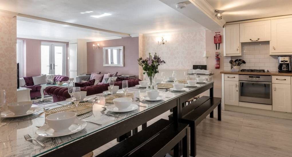 Long group dining table and open plan kitchen with purple sofa at Madame Brighton party house, 10 mins to beach.