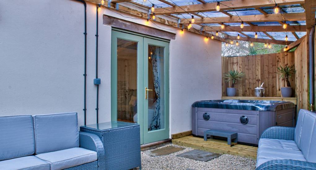 The covered outdoor patio with hot tub and grey lounge sofas at Pine Cottage, a Cotswolds hen cottage sleeping 11.