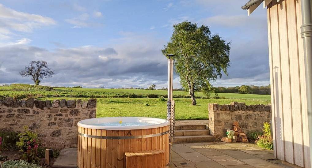 Circular wooden hot tub on stone patio with scenic field views at Heather Cottage near Edinburgh.