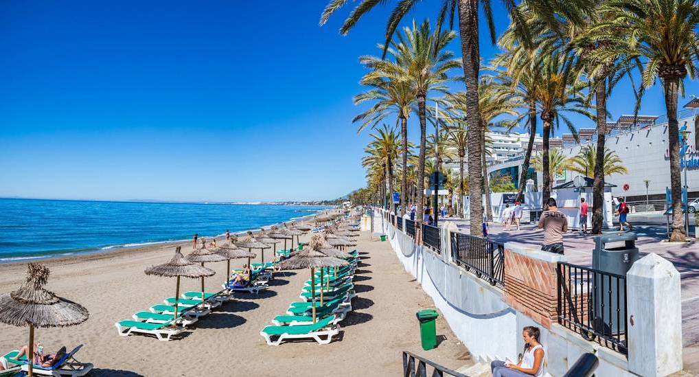 Sunny Marbella Paseo Marítimo promenade and sandy beach lined with thatched parasols, ideal for a Marbella hen do.
