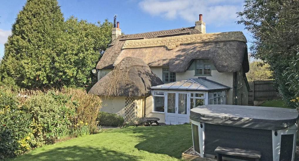 Traditional thatched cottage exterior with glass conservatory extension and outdoor hot tub on lush green lawn.