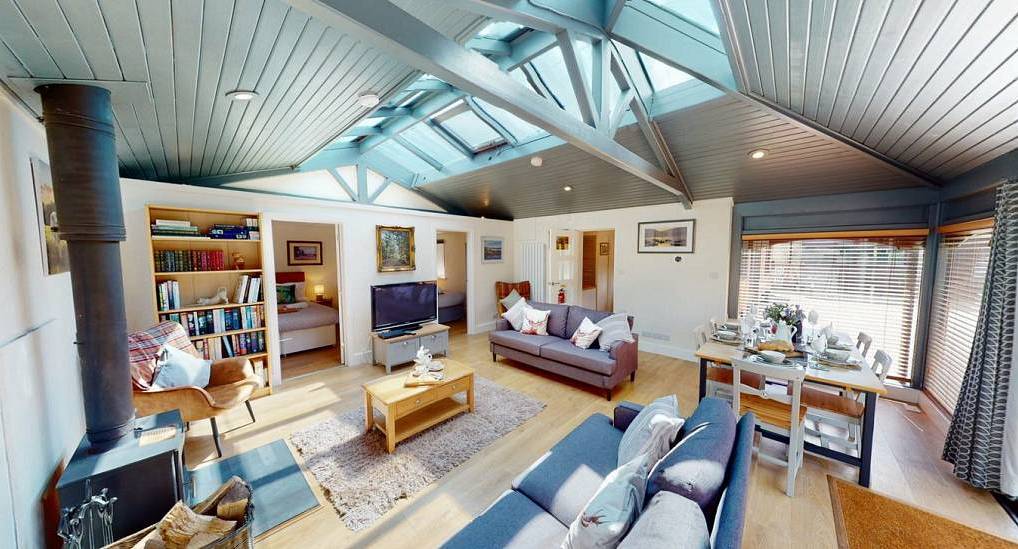 Bright open plan lounge with skylights, grey sofa, log burner stove and dining area at Stargazer lodge New Forest.