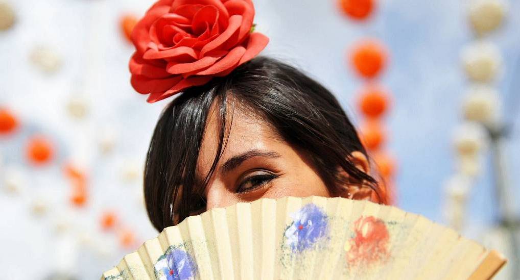Spanish woman with a hand fan and red hair flower at Feria de Abril, vibrant cultural backdrop for a Seville stag do.