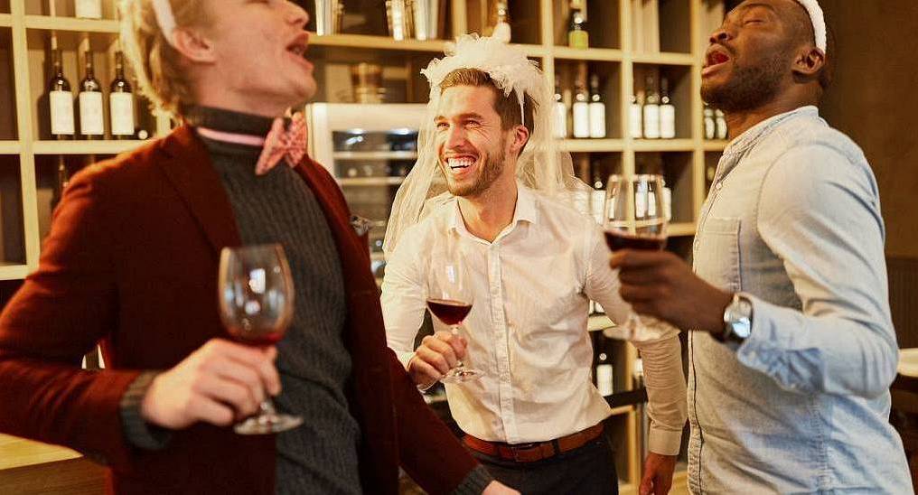 Groom wearing a veil celebrating with friends in a wine bar, lively pub crawl backdrop for a Southampton stag do.