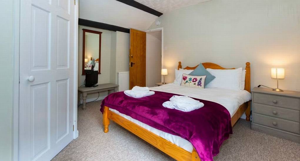 A bright double bedroom at Standing Barn Gloucestershire with a plum throw and natural wood bed frame, providing comfortable accommodation for the party house group.