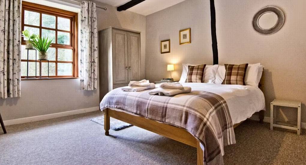 A cozy double bedroom at Standing Barn Gloucestershire featuring plaid throws and wooden ceiling beams, one of the comfortable rooms at this Gloucestershire Party House.