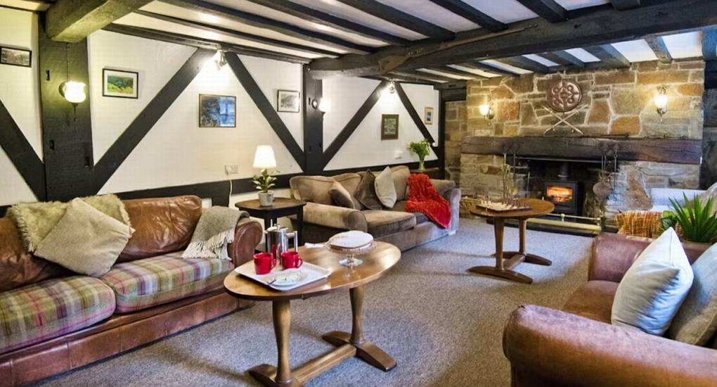 The main lounge at Standing Barn Gloucestershire, featuring comfortable sofas, exposed black and white beams, and a stone fireplace, creating a cozy Gloucestershire Party House feel.