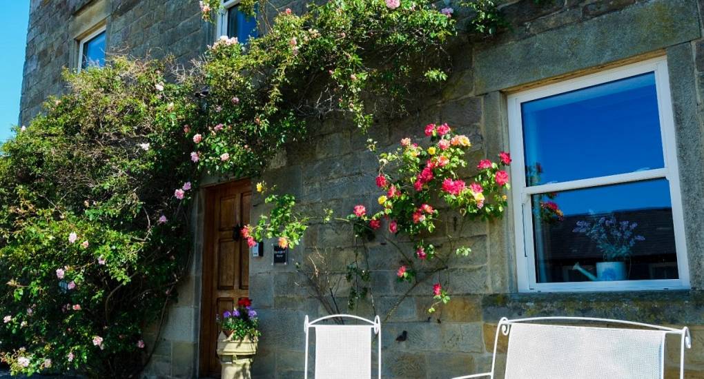 Thr front of the house at an angle with climbing plants on the walls with hanging baskets