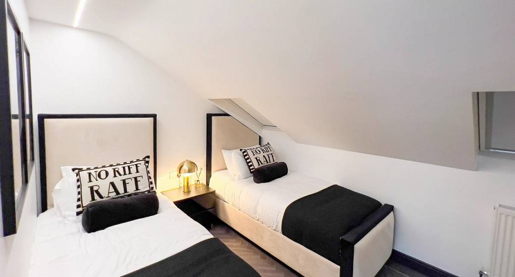 A modern, clean twin bedroom within the sloped attic space of The Plush, showcasing minimalist black-and-white decor and fun 'No Riff Raff' throw pillows.