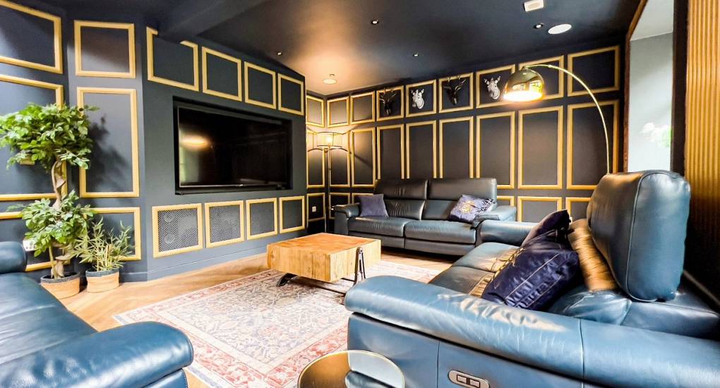 The cinematic lounge at The Plush, a luxury Newcastle hen do house, featuring dramatic gold-trimmed paneling, a large integrated flat-screen TV, and plush leather sofas for group socializing.