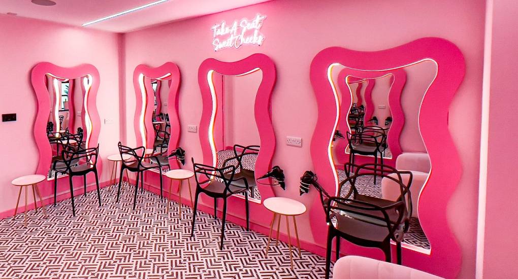 A close-up of the vibrant pink dressing room at The Plush, showcasing the 'Take A Seat Sweet Cheeks' neon sign and glam vanity mirrors perfect for hen group prep.