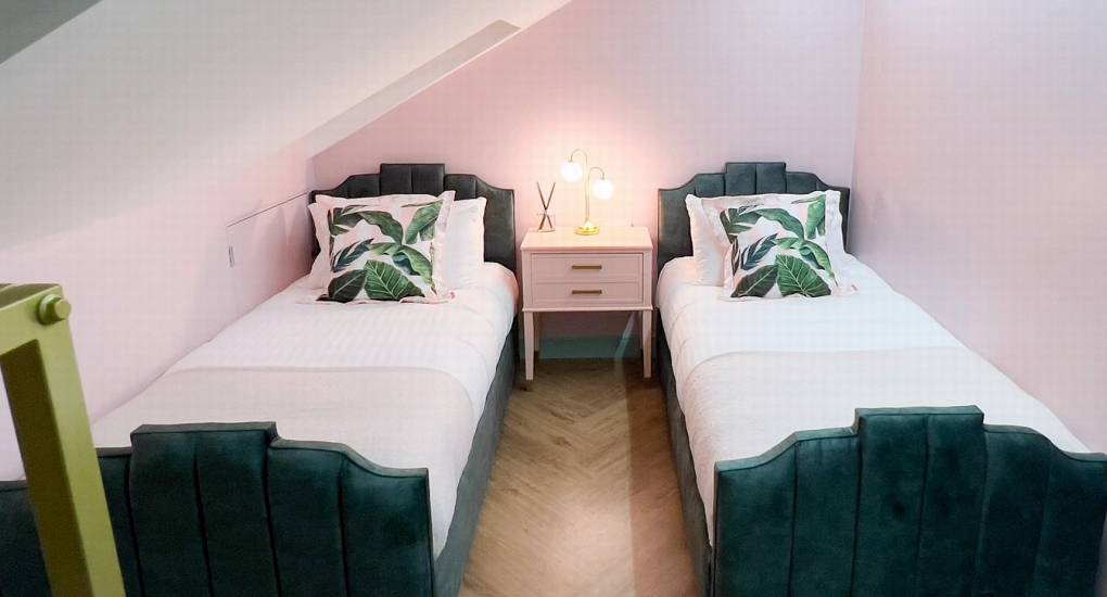 A chic twin bedroom at The Plush with softly lit walls, featuring decorative fluted teal headboards, tropical leaf pillows, and a glamorous vanity-style nightstand.