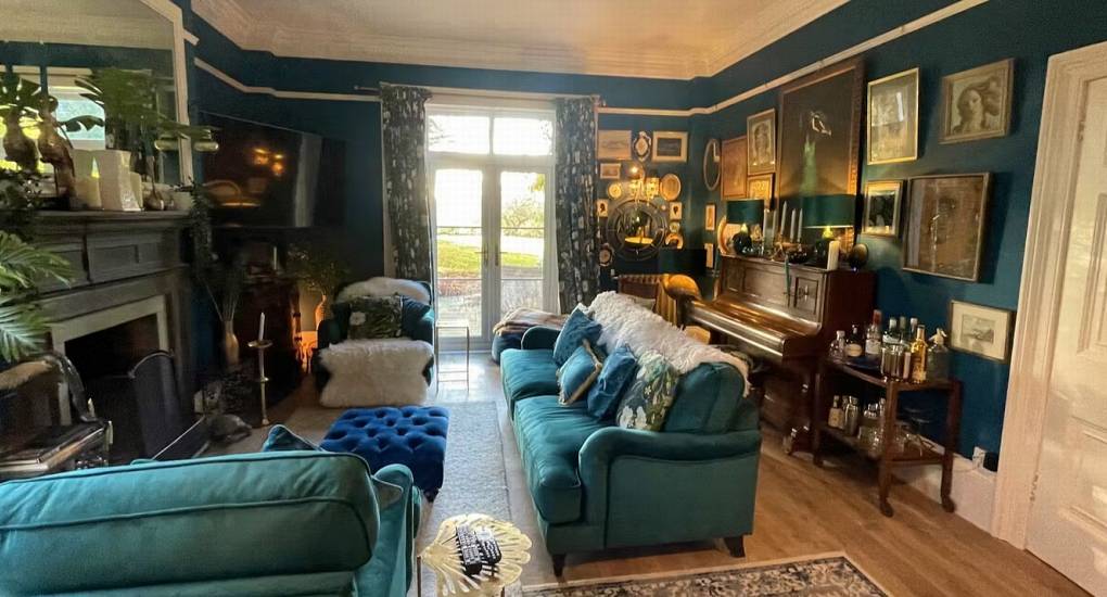  A view of the beautifully decorated lounge and drawing room at The Sash House, a cozy space with two large blue sofas, a piano, and a large fireplace, perfect for a hen do cottage.