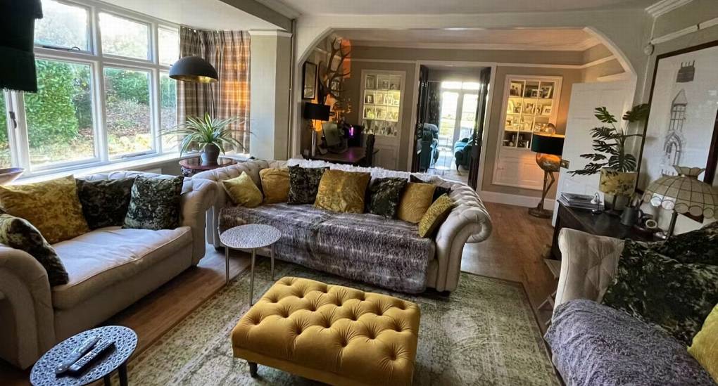The colorful and eclectic lounge area at The Sash House, with a large chesterfield-style sofa, a mustard yellow ottoman, and large windows looking out onto the grounds of this Herefordshire hen do cottage.
