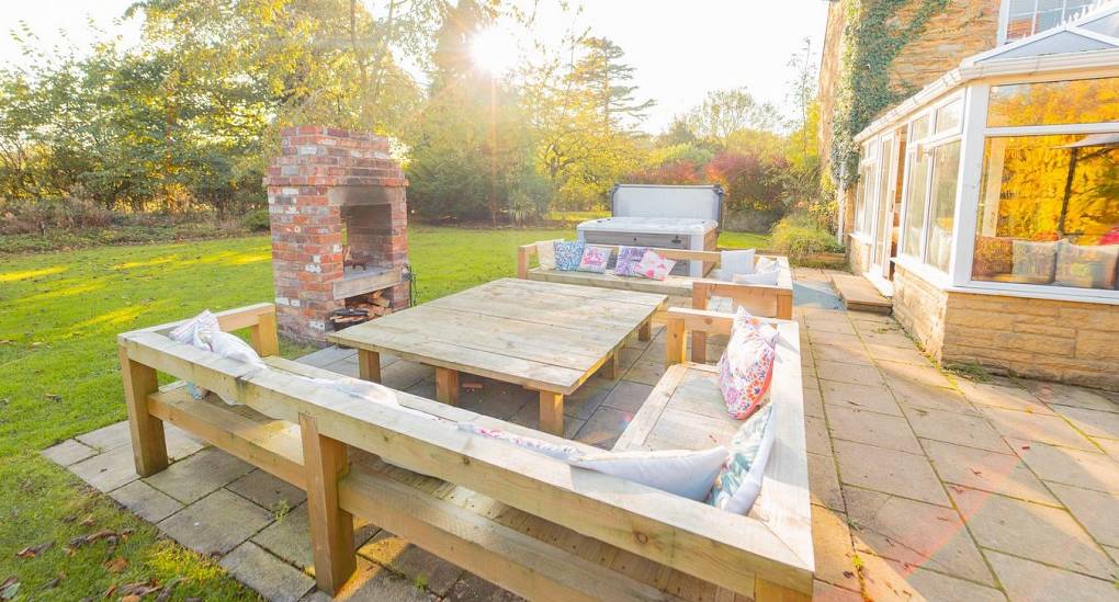 The outdoor social oasis at Tranquil Escape, a rural Newcastle hen do house, featuring a brick-built fire pit, large rustic wooden seating, and a modern hot tub set in private grounds.